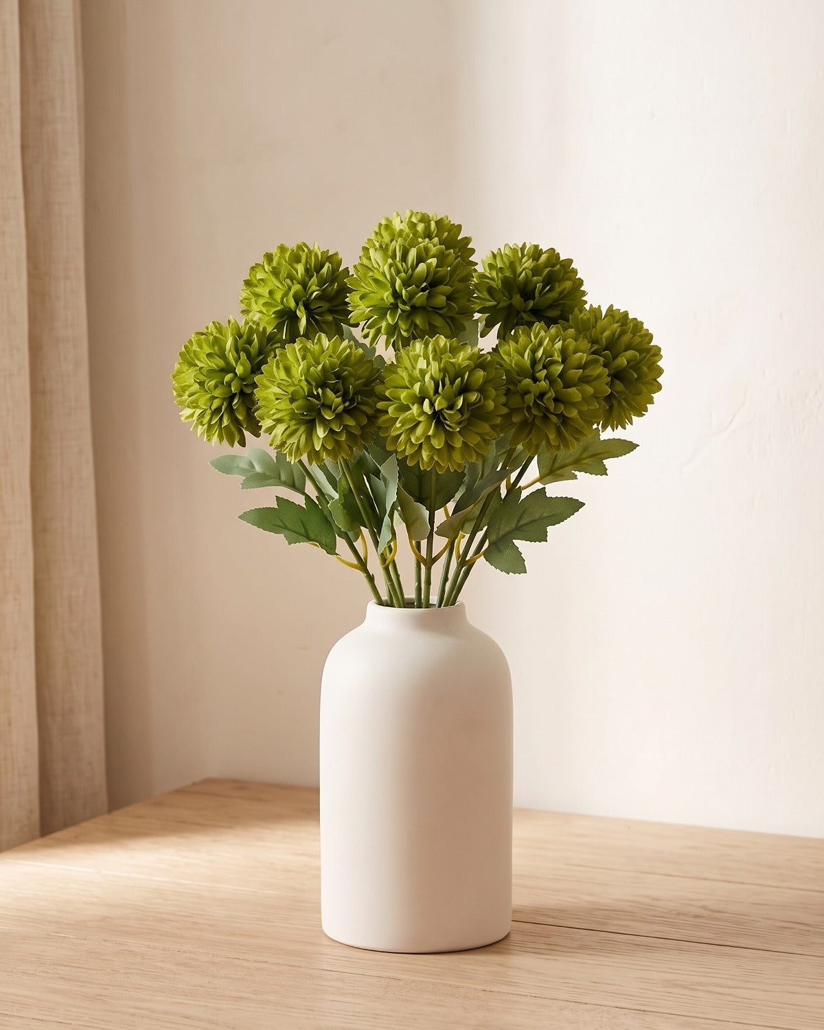 Olive Green artificial pompom chrysanthemums standing in a vase on a pale oak table; no vase is included