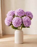 Light Purple artificial hydrangea flowers standing in a plain off-white vase on a pale oak table; no vase is included