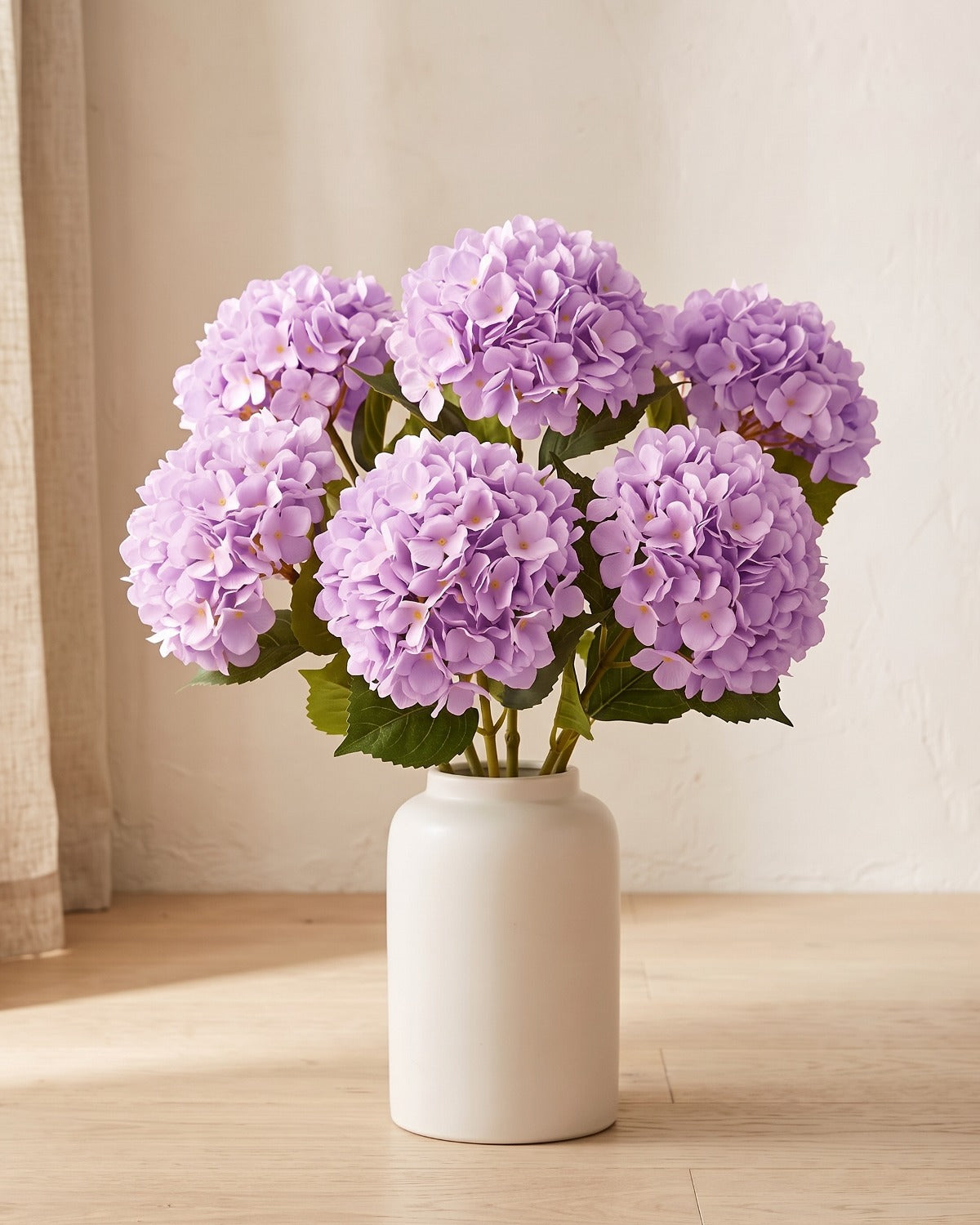 Light Purple artificial hydrangea flowers standing in a plain off-white vase on a pale oak table; no vase is included