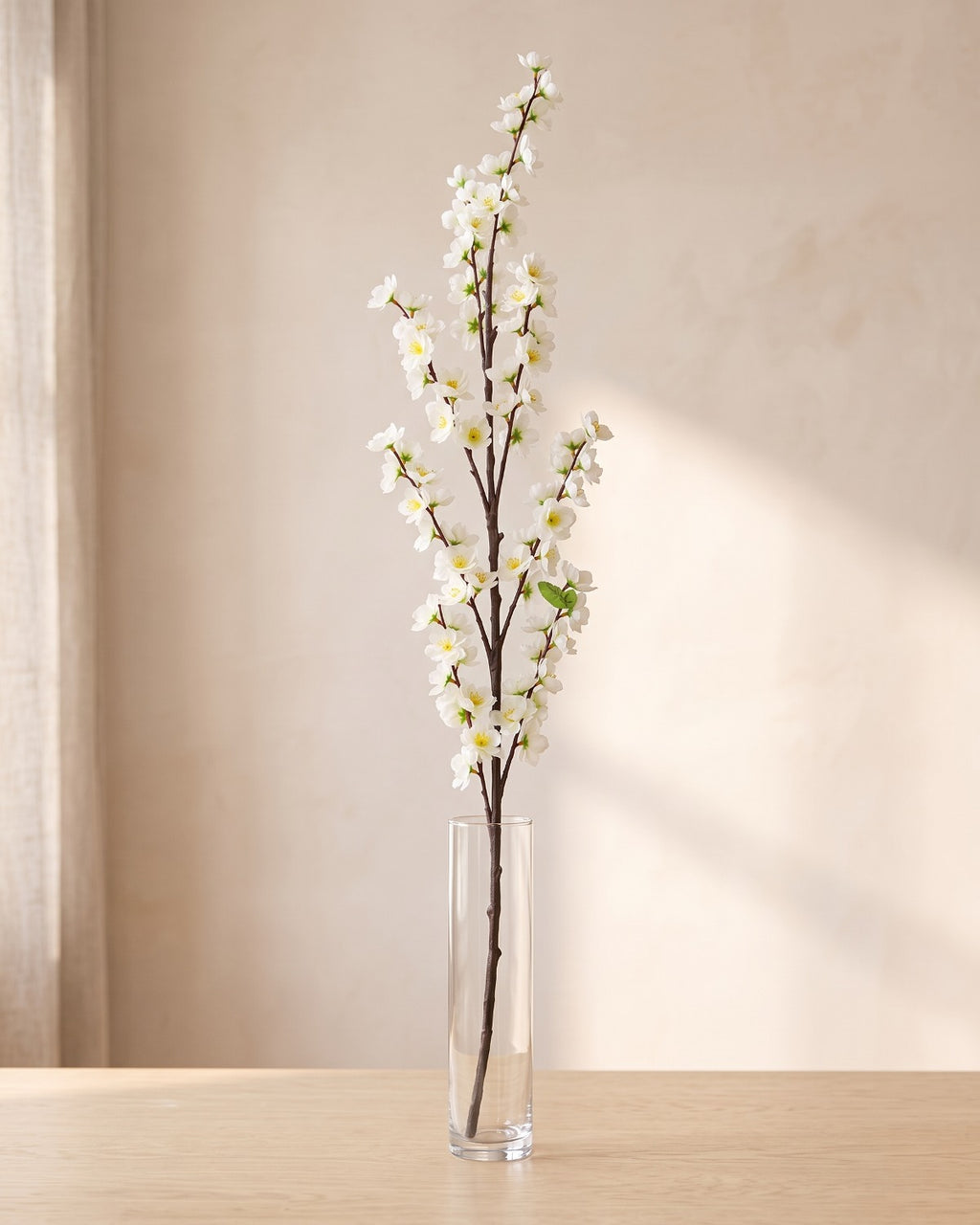 White artificial peach blossom branch standing in a tall glass vase on a pale oak table; no vase is included