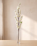 White artificial peach blossom branch standing in a tall glass vase on a pale oak table; no vase is included