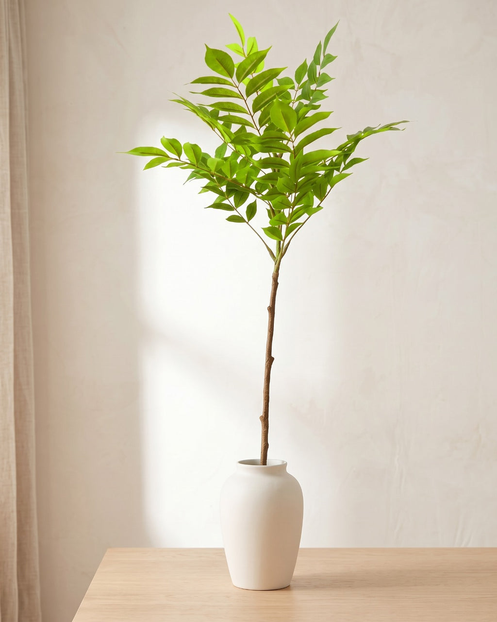 Feather leaf artificial tree branch standing in a plain off-white vase on a pale oak table; no vase is included