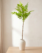 Feather leaf artificial tree branch standing in a plain off-white vase on a pale oak table; no vase is included