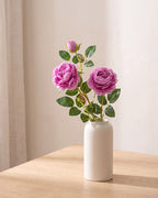 Purple artificial rose branch standing in a plain off-white vase on a pale oak table; each order is one branch, and no vase is included