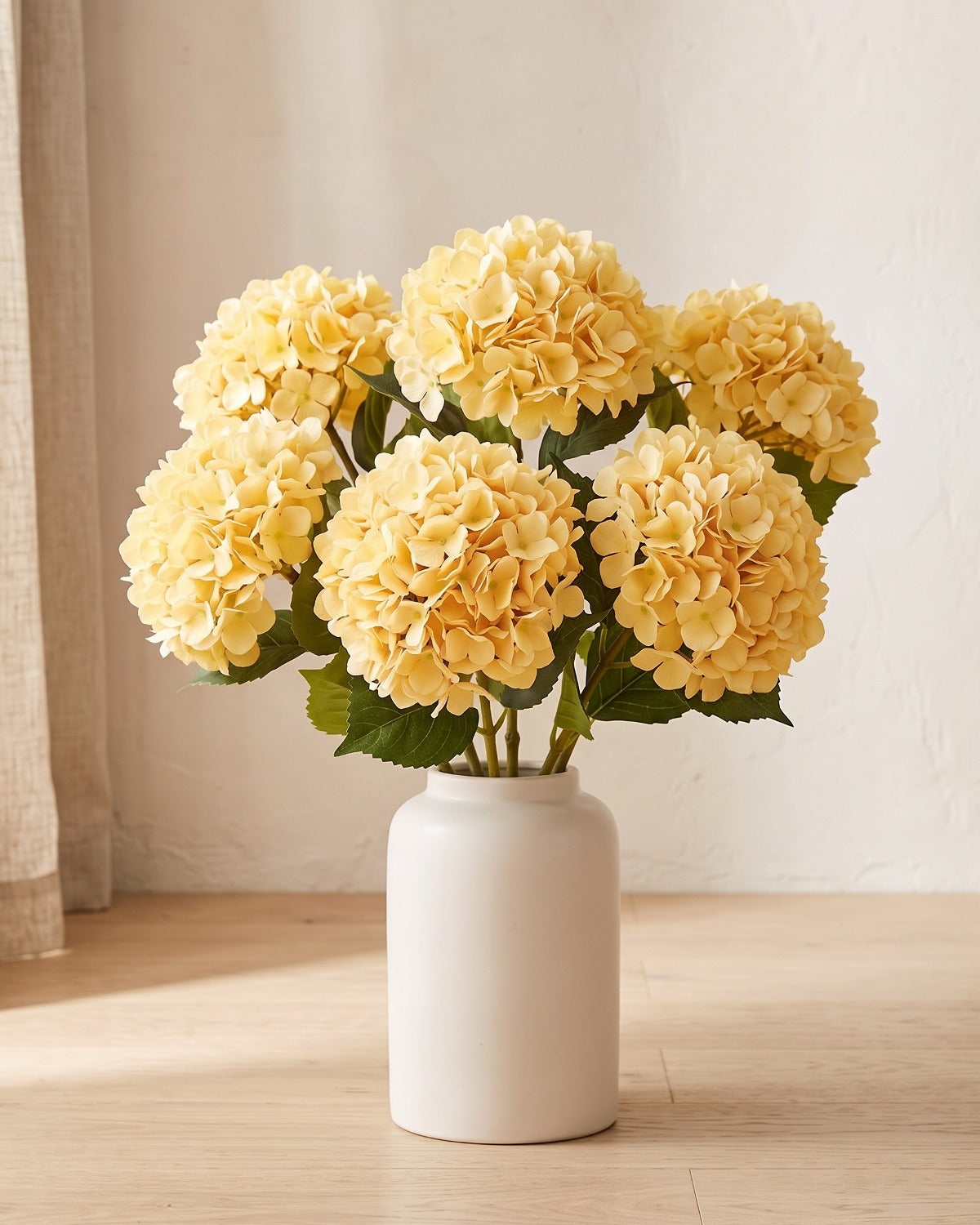 Yellow artificial hydrangea flowers standing in a plain off-white vase on a pale oak table; no vase is included
