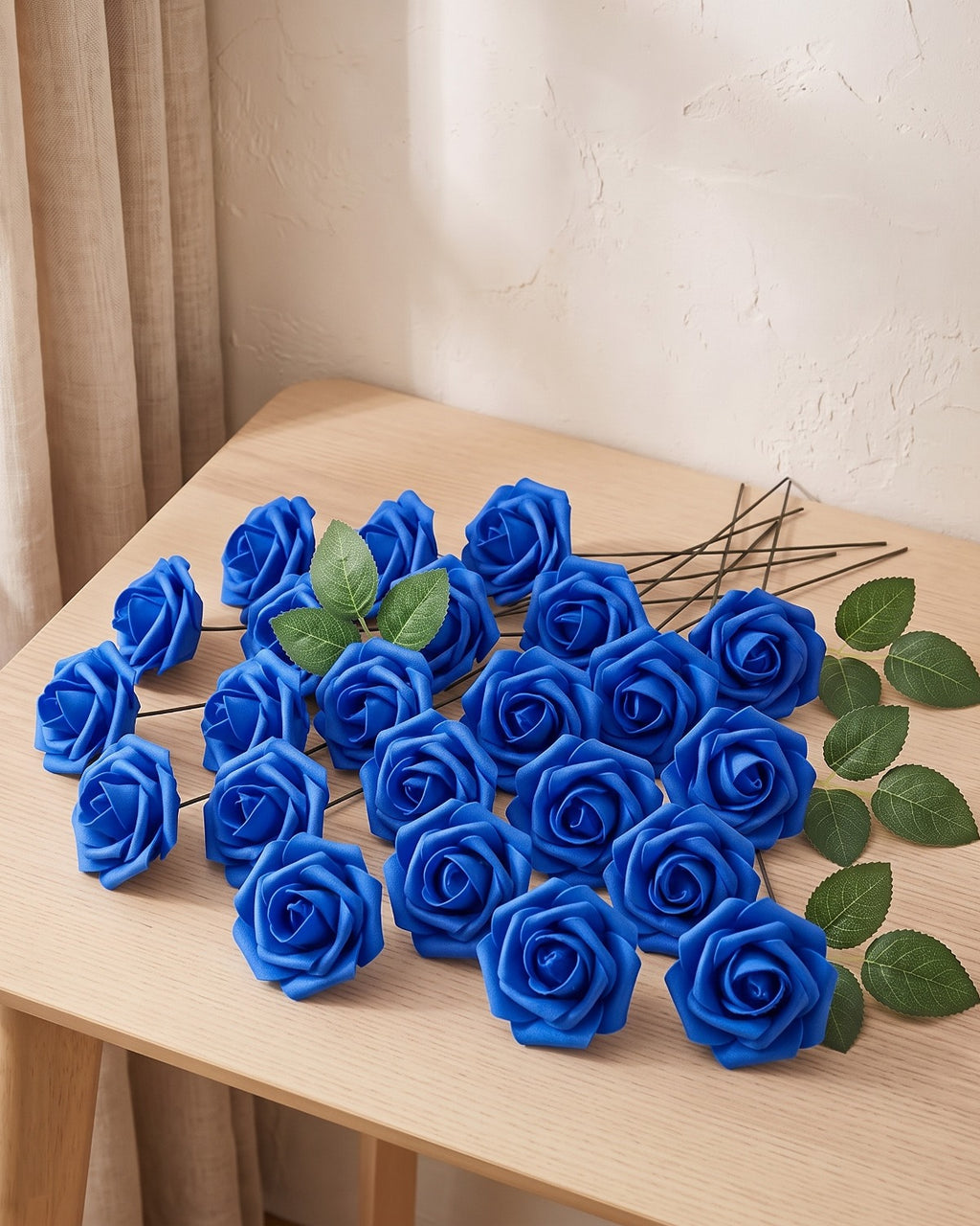 Twenty-five royal blue foam rose heads on wire stems with green leaf sprays, laid out on a pale oak table