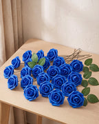 Twenty-five royal blue foam rose heads on wire stems with green leaf sprays, laid out on a pale oak table