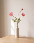 A pink artificial poppy stem, one stem with two open flowers and a bud, in a plain off-white bud vase on a pale oak table; vase not included