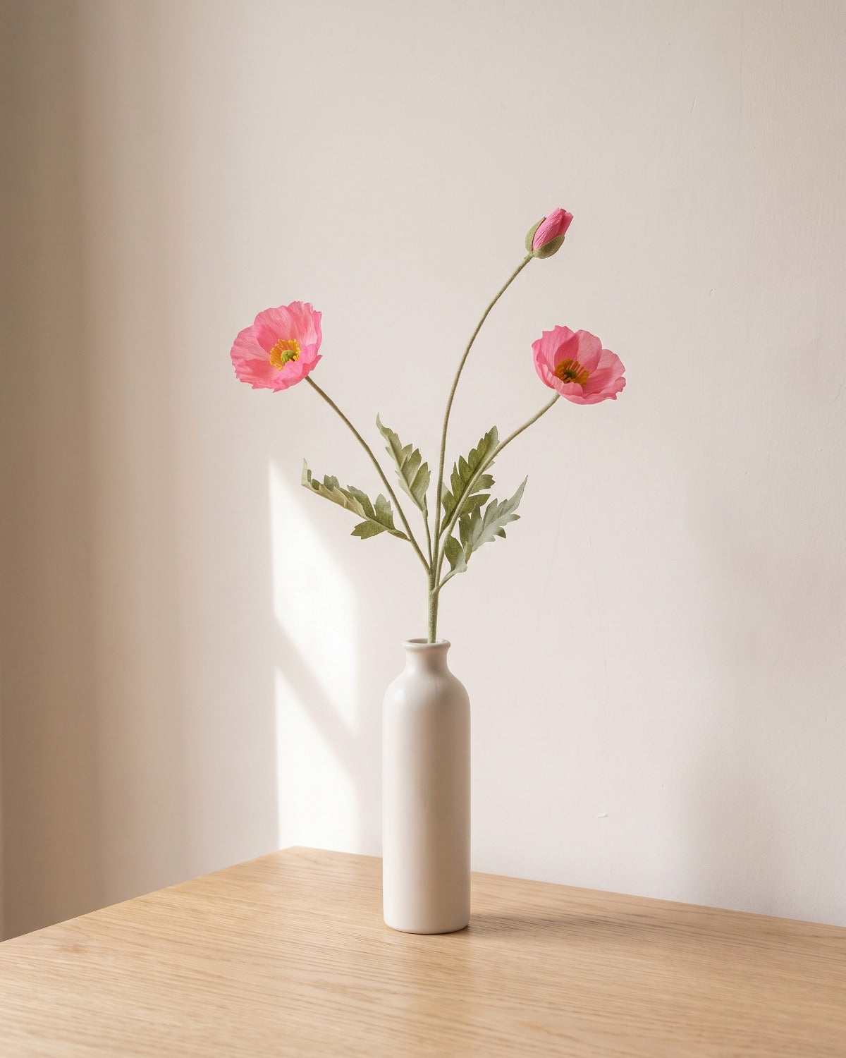 A pink artificial poppy stem, one stem with two open flowers and a bud, in a plain off-white bud vase on a pale oak table; vase not included