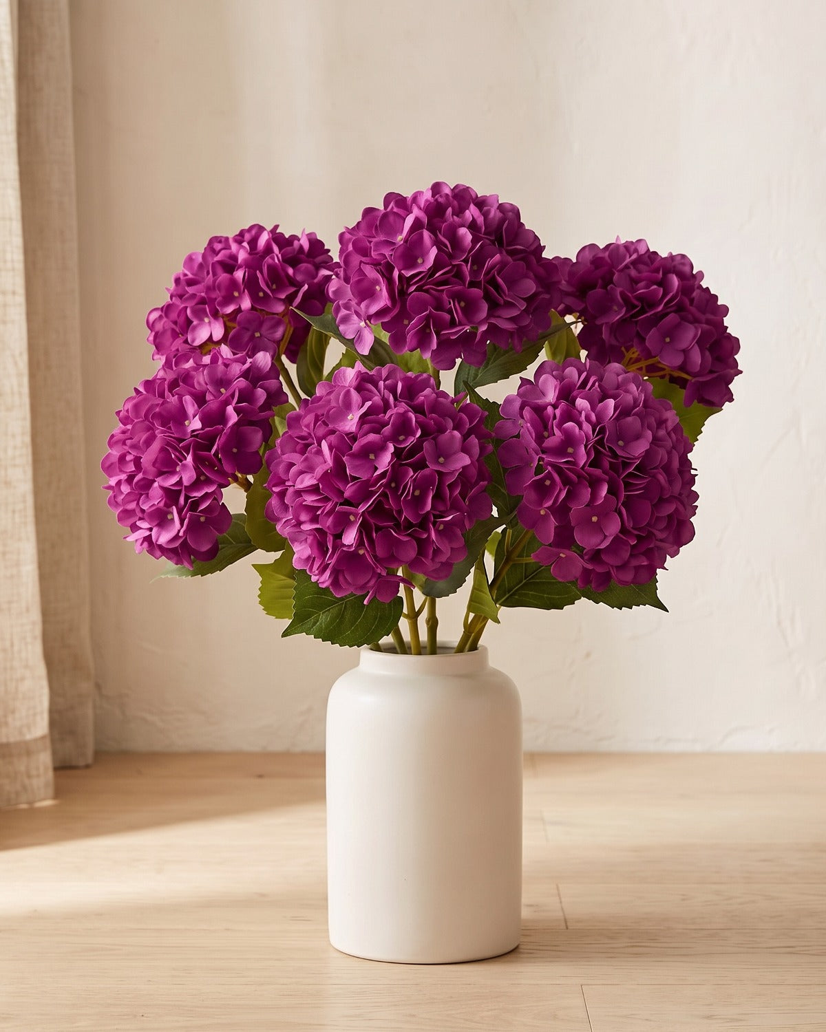 Purple artificial hydrangea flowers standing in a plain off-white vase on a pale oak table; no vase is included