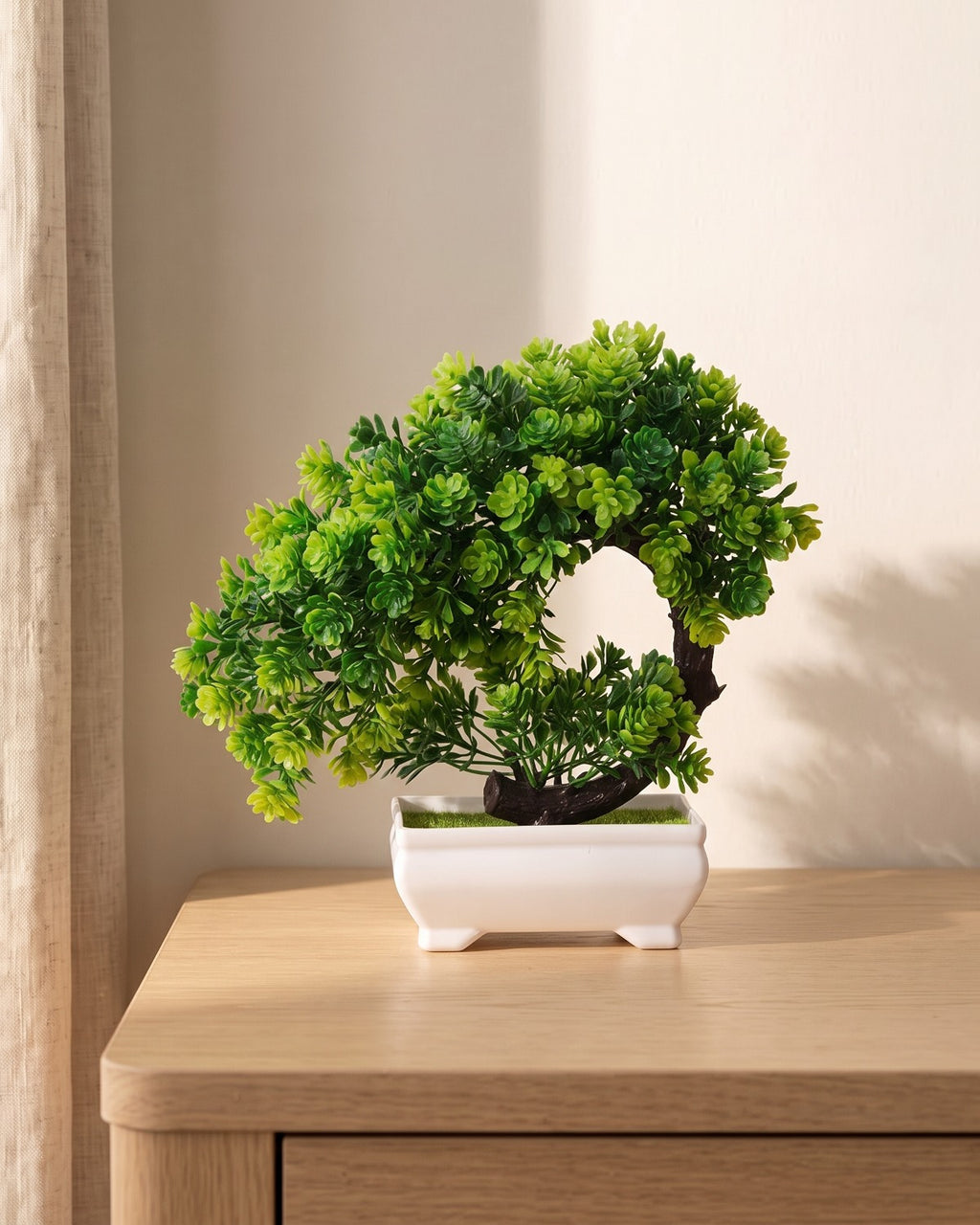 A small artificial green bonsai with a cascading trunk, the foliage falling below the rim, in the white ceramic tray it comes in, on a pale oak table