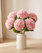 Pink artificial hydrangea flowers standing in a plain off-white vase on a pale oak table; no vase is included