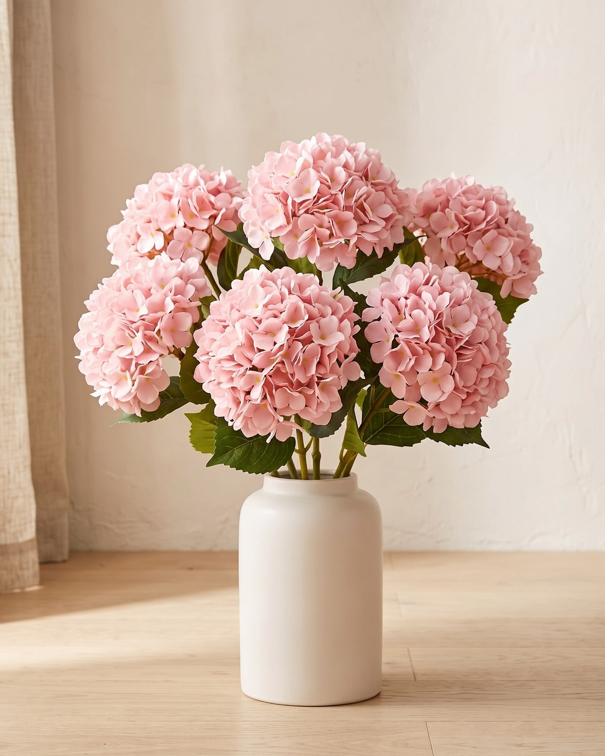 Pink artificial hydrangea flowers standing in a plain off-white vase on a pale oak table; no vase is included