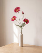 A dark pink artificial poppy stem, one stem with two open flowers and a bud, in a plain off-white bud vase on a pale oak table; vase not included