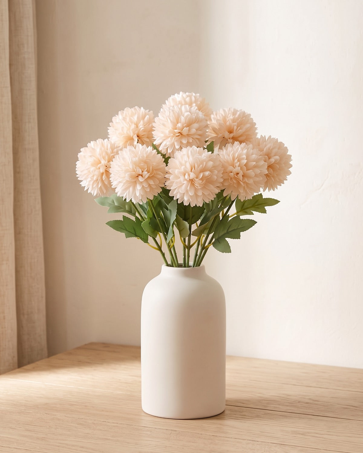 Champagne artificial pompom chrysanthemums standing in a vase on a pale oak table; no vase is included