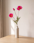 A fuchsia artificial poppy stem, one stem with two open flowers and a bud, in a plain off-white bud vase on a pale oak table; vase not included