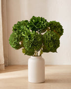 Green artificial hydrangea flowers standing in a plain off-white vase on a pale oak table; no vase is included