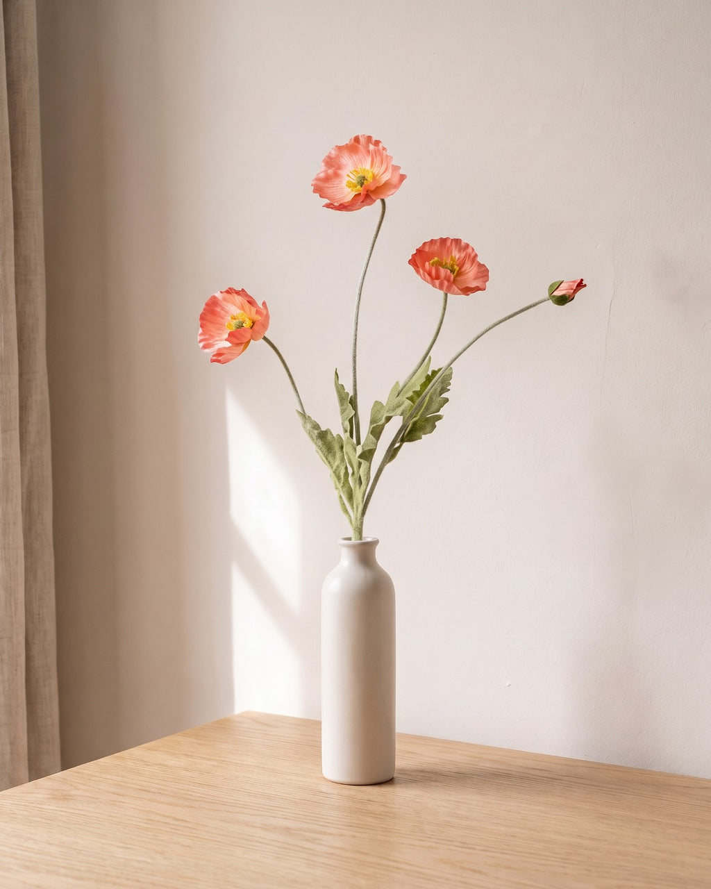 A coral pink artificial poppy stem, one stem with two open flowers and a bud, in a plain off-white bud vase on a pale oak table; vase not included