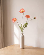 A coral pink artificial poppy stem, one stem with two open flowers and a bud, in a plain off-white bud vase on a pale oak table; vase not included