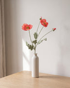 A dark coral artificial poppy stem, one stem with two open flowers and a bud, in a plain off-white bud vase on a pale oak table; vase not included