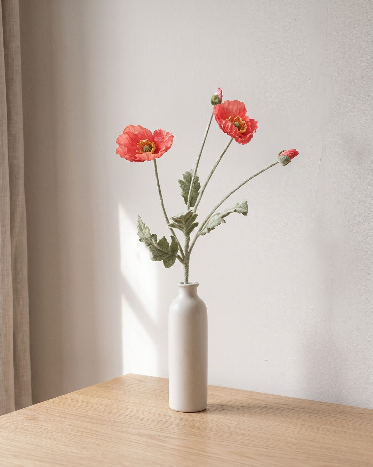 A dark coral artificial poppy stem, one stem with two open flowers and a bud, in a plain off-white bud vase on a pale oak table; vase not included