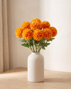 Orange artificial pompom chrysanthemums standing in a vase on a pale oak table; no vase is included