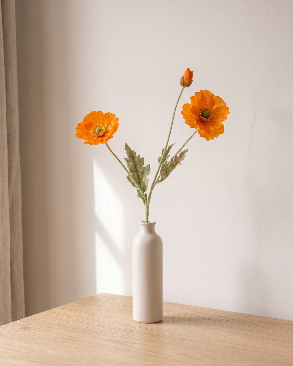 A orange artificial poppy stem, one stem with two open flowers and a bud, in a plain off-white bud vase on a pale oak table; vase not included