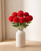 Red artificial pompom chrysanthemums standing in a vase on a pale oak table; no vase is included