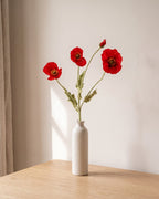 A red orange artificial poppy stem, one stem with two open flowers and a bud, in a plain off-white bud vase on a pale oak table; vase not included