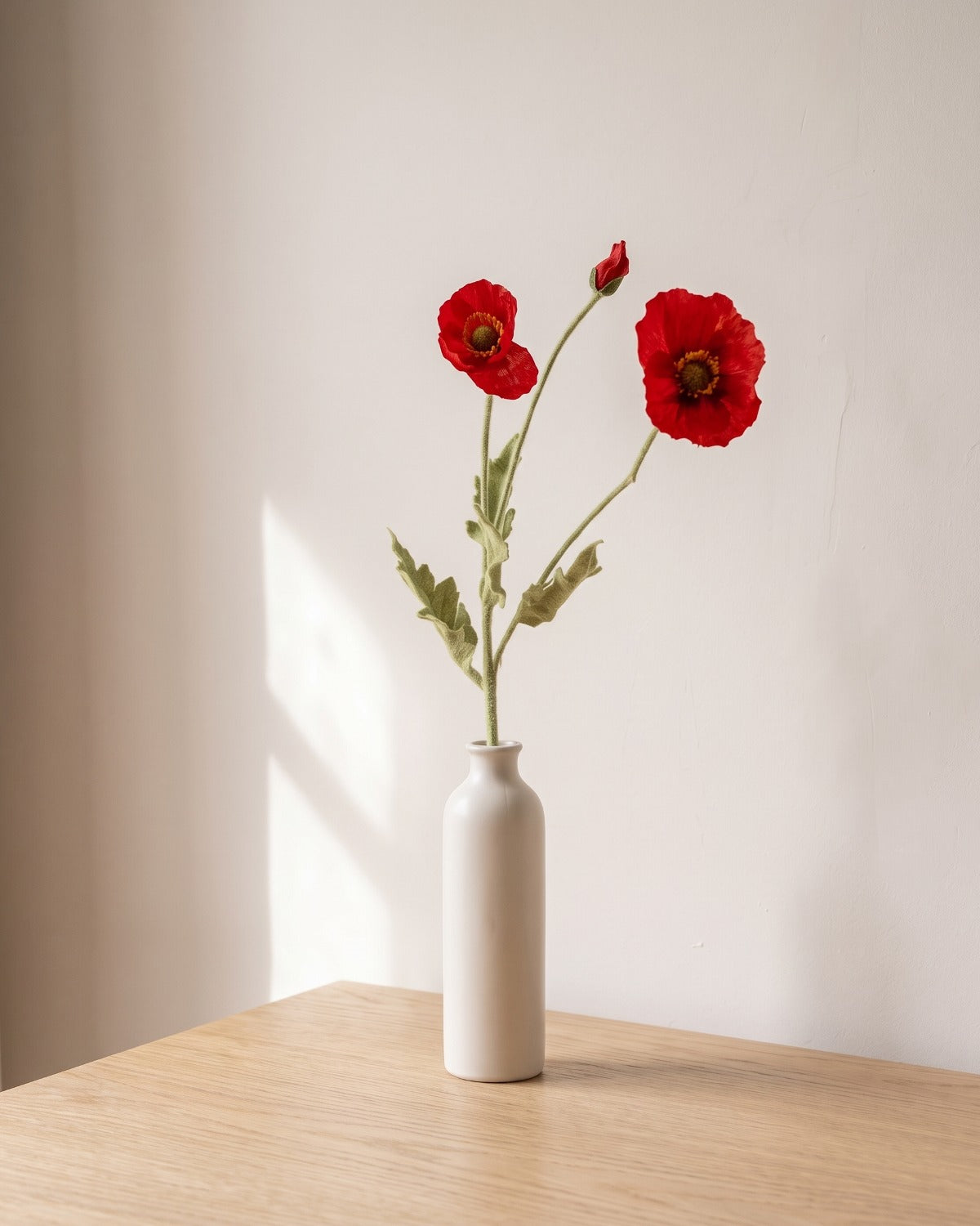 A red artificial poppy stem, one stem with two open flowers and a bud, in a plain off-white bud vase on a pale oak table; vase not included