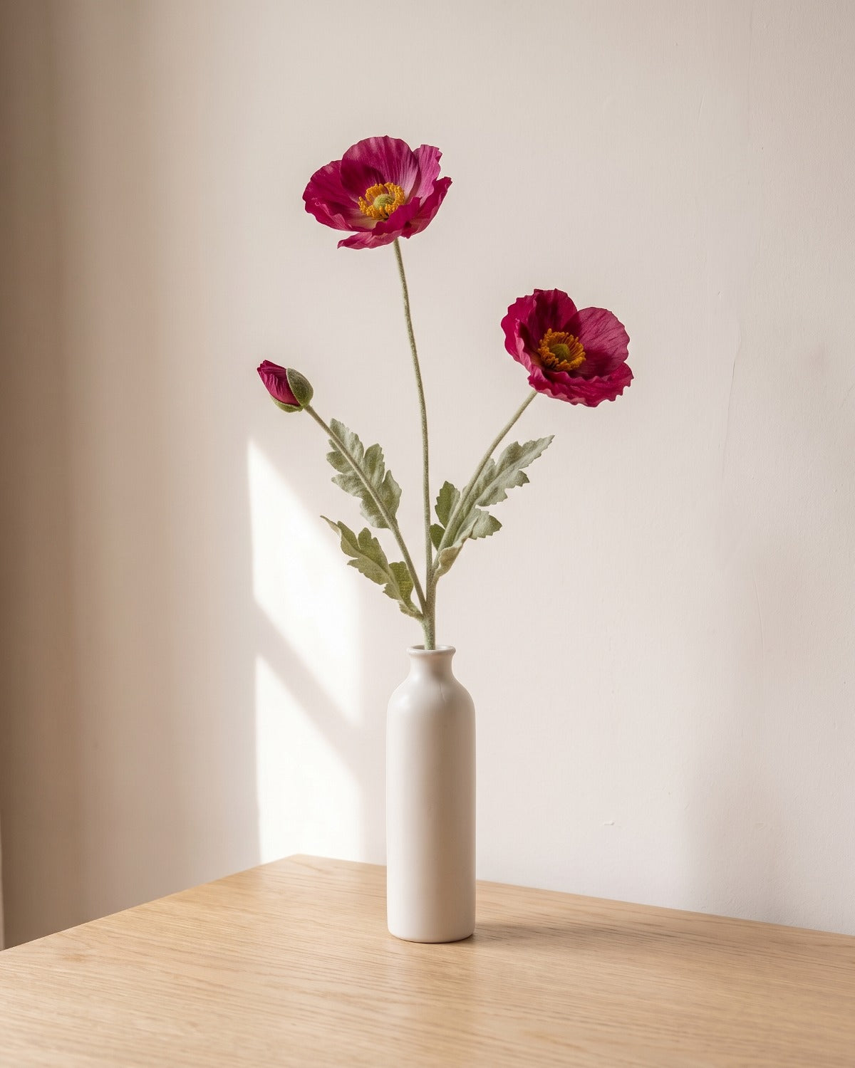 A purple red artificial poppy stem, one stem with two open flowers and a bud, in a plain off-white bud vase on a pale oak table; vase not included