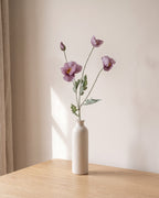 A mauve artificial poppy stem, one stem with two open flowers and a bud, in a plain off-white bud vase on a pale oak table; vase not included