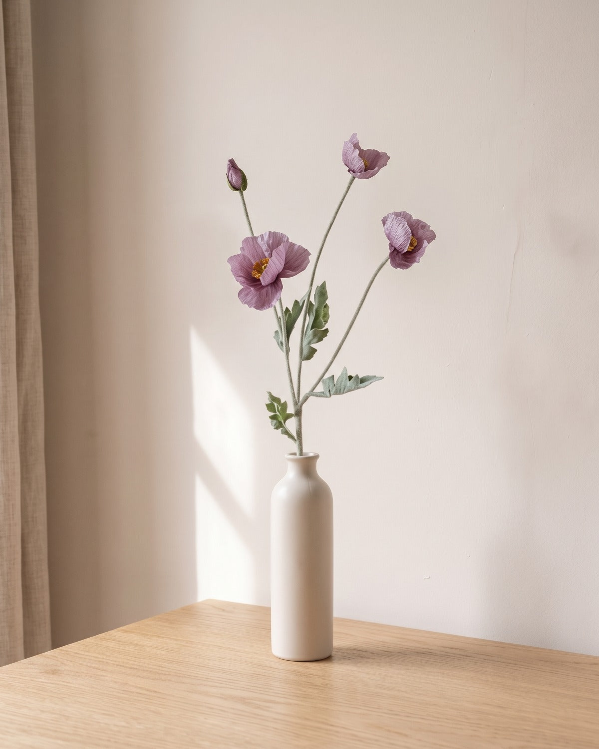 A mauve artificial poppy stem, one stem with two open flowers and a bud, in a plain off-white bud vase on a pale oak table; vase not included