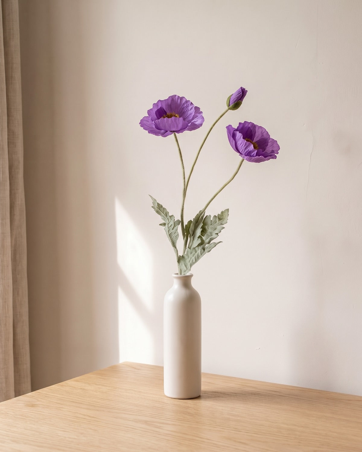 A purple artificial poppy stem, one stem with two open flowers and a bud, in a plain off-white bud vase on a pale oak table; vase not included