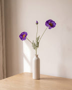 A dark purple artificial poppy stem, one stem with two open flowers and a bud, in a plain off-white bud vase on a pale oak table; vase not included