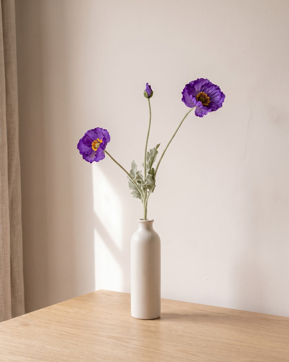 A dark purple artificial poppy stem, one stem with two open flowers and a bud, in a plain off-white bud vase on a pale oak table; vase not included