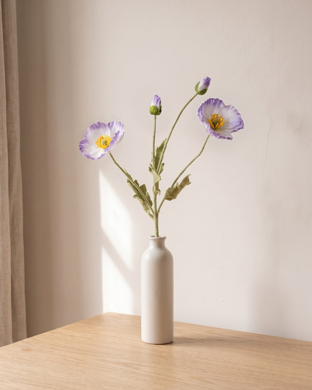 A purple white artificial poppy stem, one stem with two open flowers and a bud, in a plain off-white bud vase on a pale oak table; vase not included
