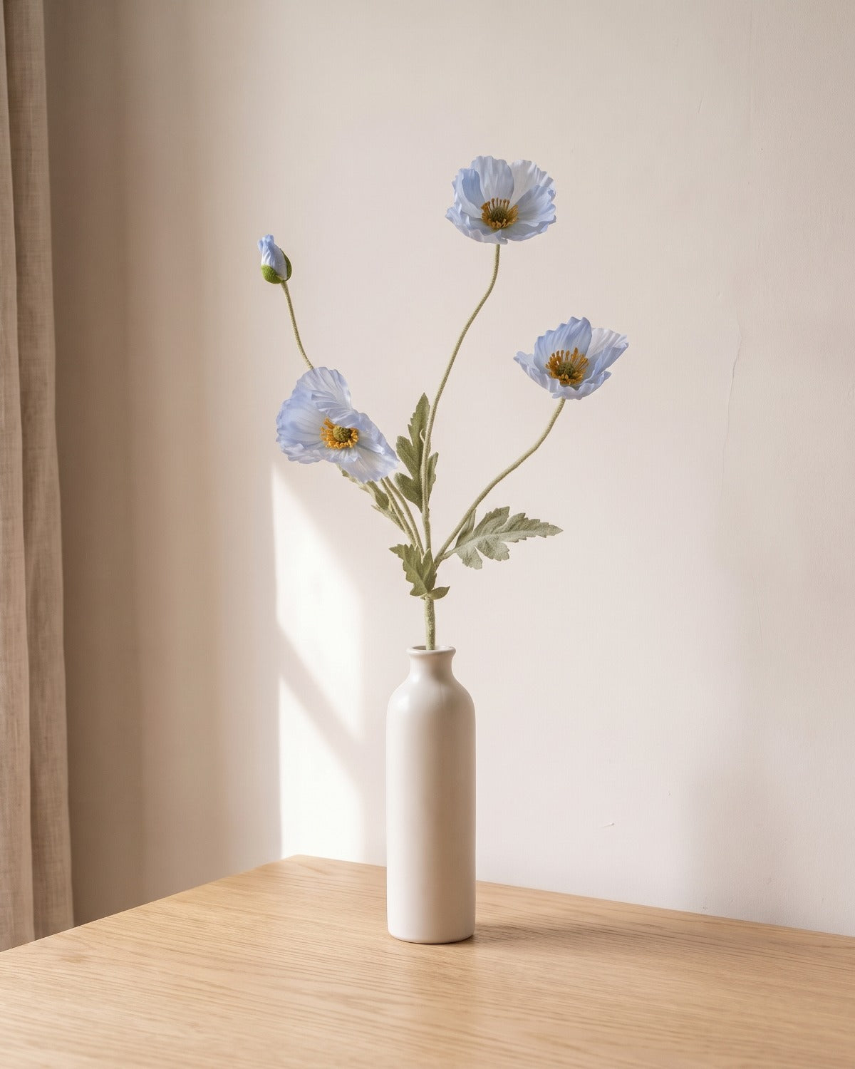 A light blue artificial poppy stem, one stem with two open flowers and a bud, in a plain off-white bud vase on a pale oak table; vase not included