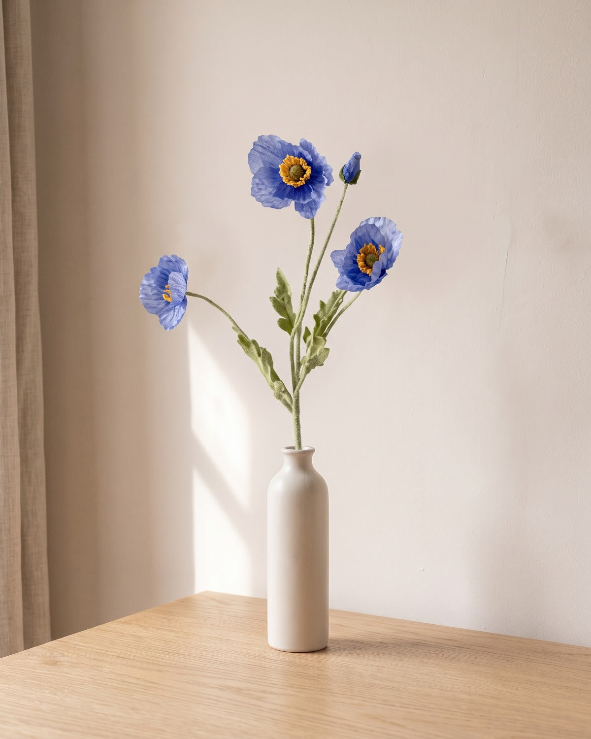 A blue artificial poppy stem, one stem with two open flowers and a bud, in a plain off-white bud vase on a pale oak table; vase not included
