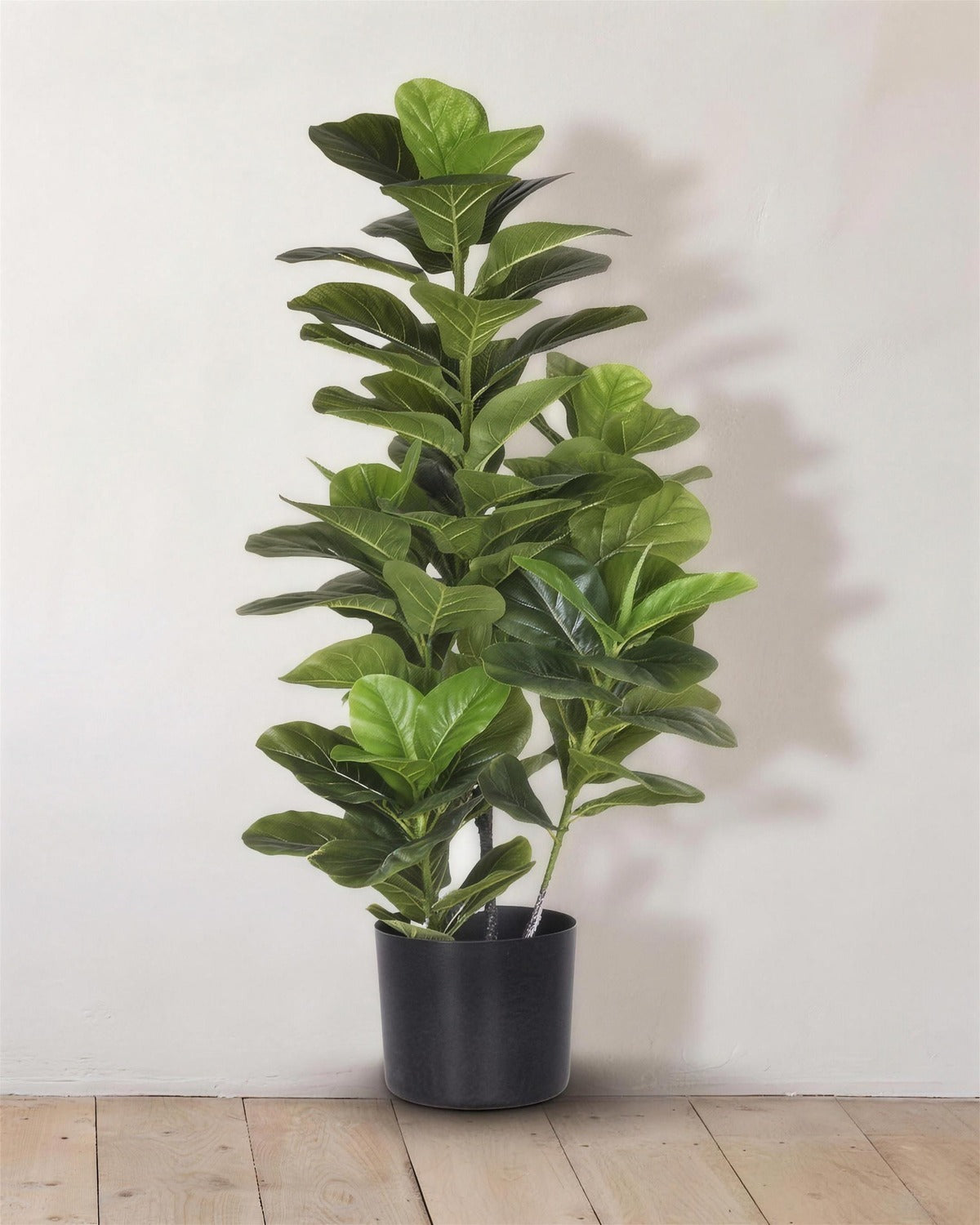 An artificial fiddle leaf fig tree in the 39.4 in and 47.2 in sizes, in its black nursery pot against a warm white wall
