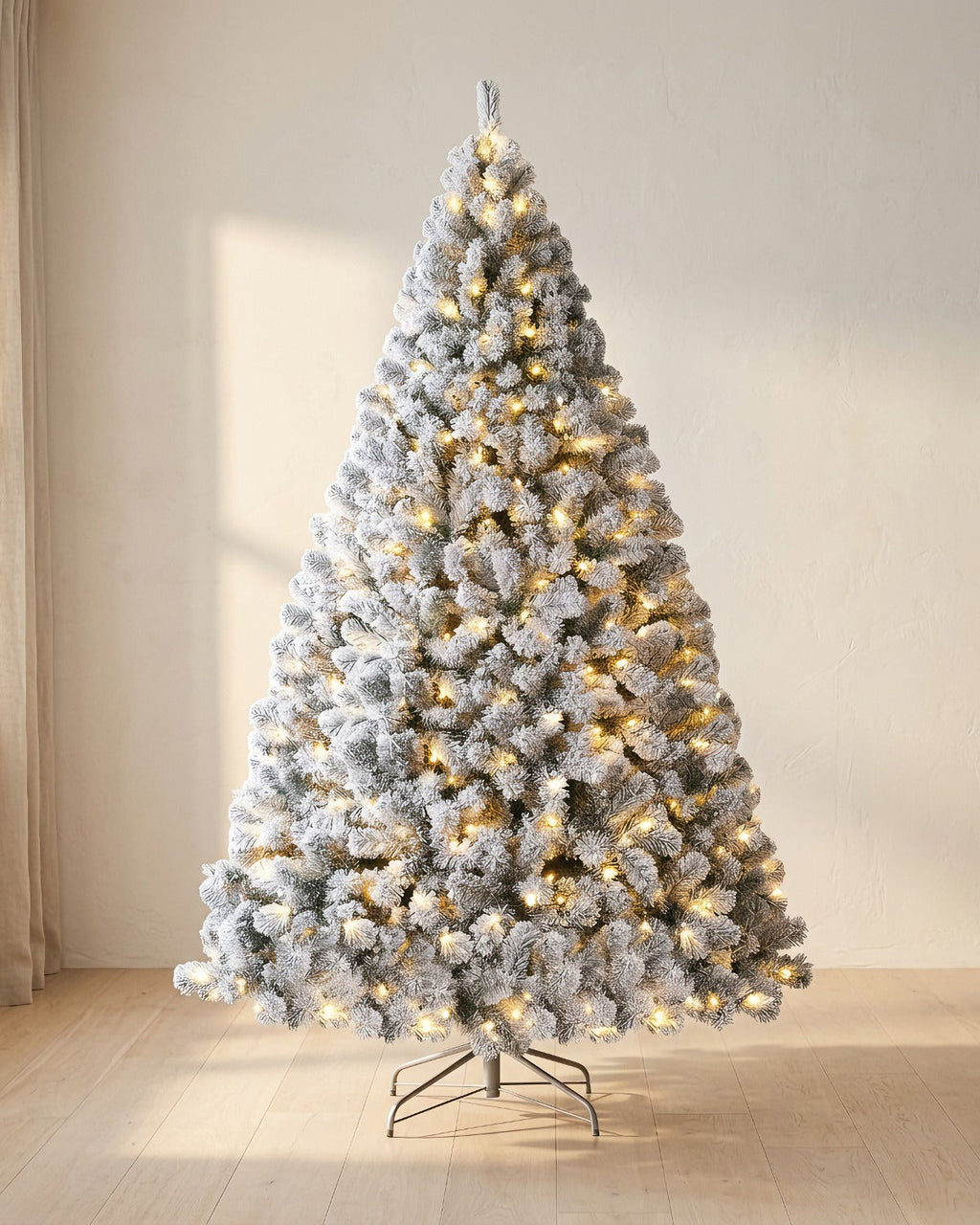 Pre-lit snow flocked Christmas tree with its warm white lights on, beside a linen curtain
