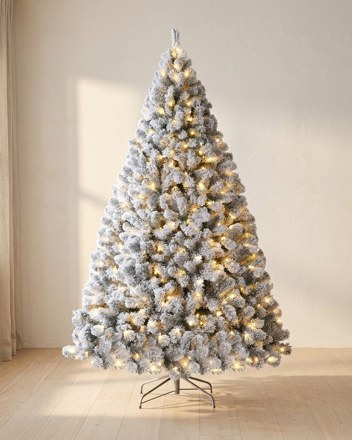 Pre-lit snow flocked Christmas tree with its warm white lights on, beside a linen curtain