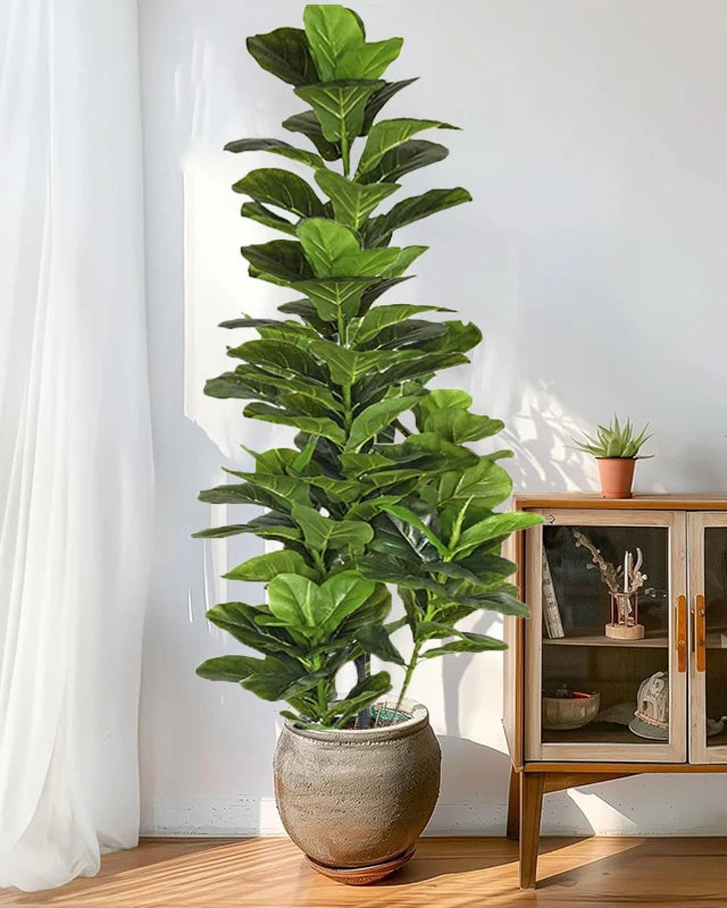 Artificial fiddle leaf fig tree in a stone pot beside a sheer curtain and a cabinet