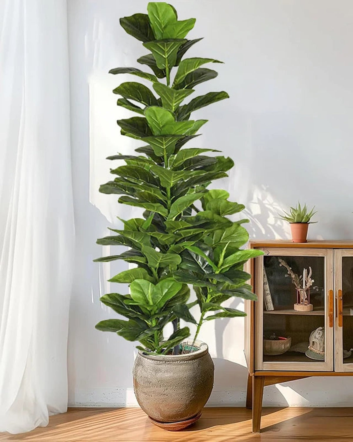 Artificial fiddle leaf fig tree in a stone pot beside a sheer curtain and a cabinet
