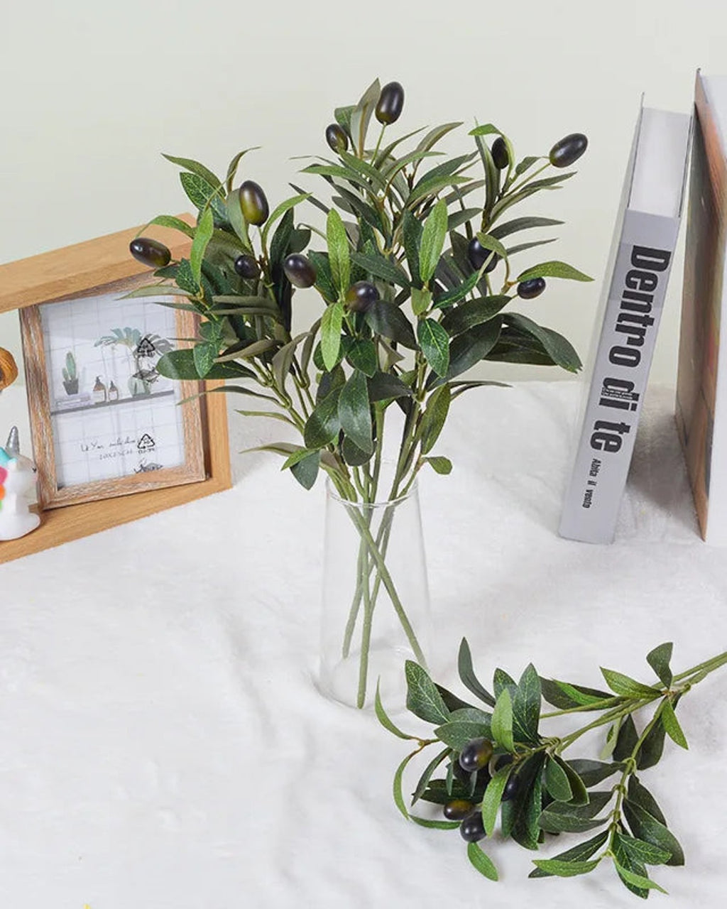 Artificial olive branches in a glass vase beside picture frames and books
