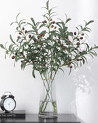 Artificial olive branches in a tall glass vase beside an alarm clock