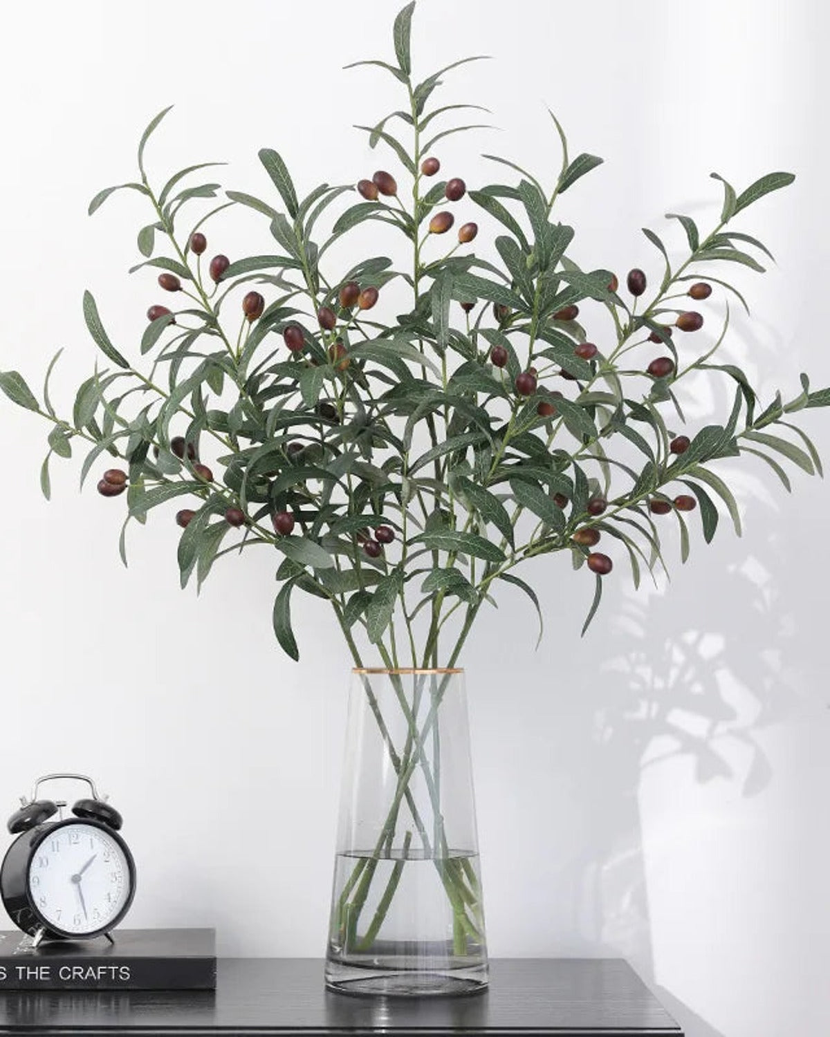 Artificial olive branches in a tall glass vase beside an alarm clock