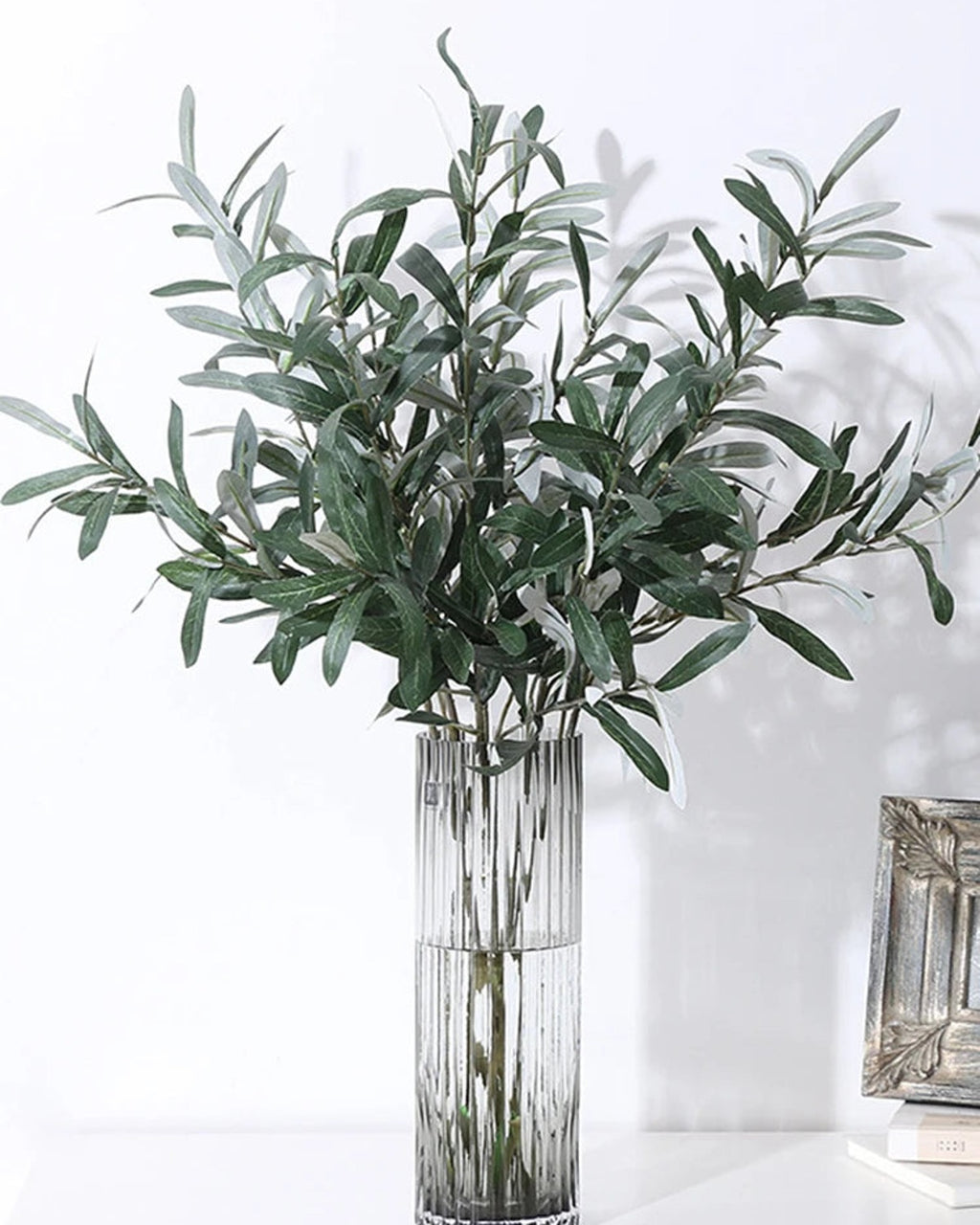 Artificial olive branches in a ribbed glass vase on a white table