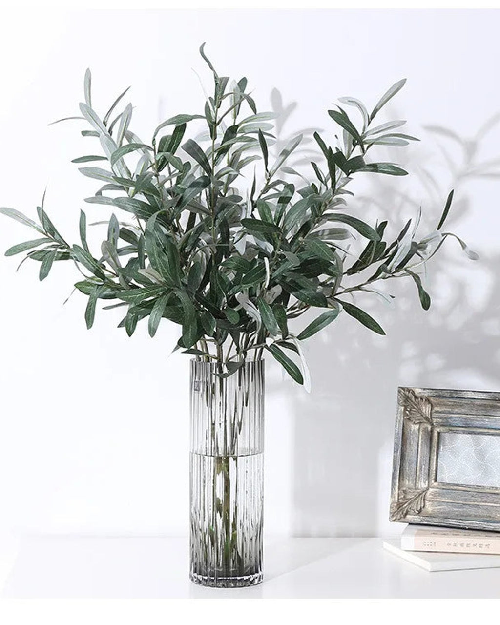 Artificial olive branches in a ribbed glass vase beside a picture frame