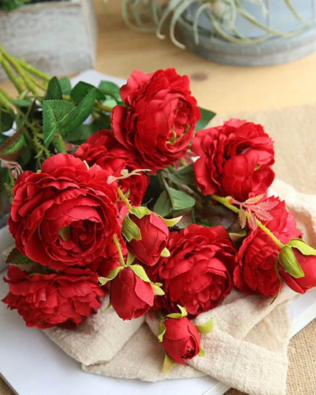 Red artificial rose branches laid on linen, showing the cupped many petalled blooms; each order is one branch